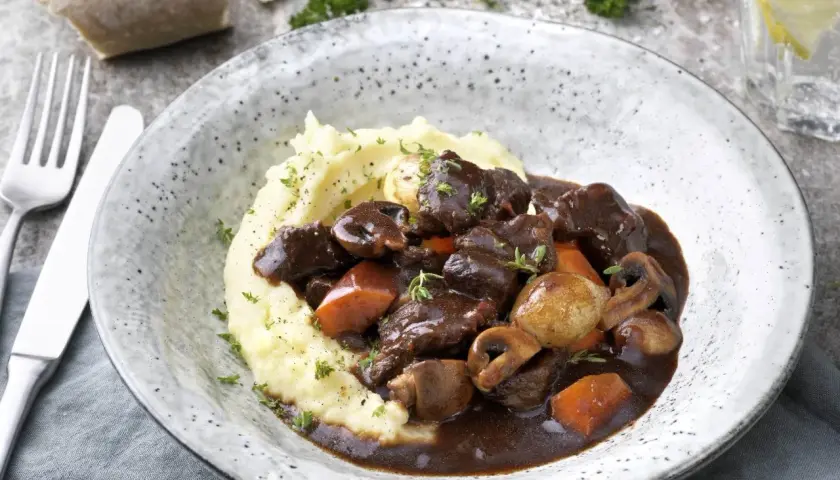 En mustig boeuf bourguignon med potatismos, champinjoner och morötter. Serveras i en rustik skål med en bit bröd och ett glas vatten.
