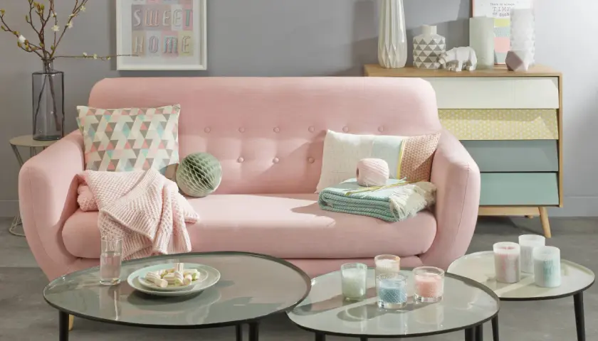 Sofá rosa pastel con cojines y mantas, mesas de centro con velas y malvaviscos, y un mueble de cajones de colores.