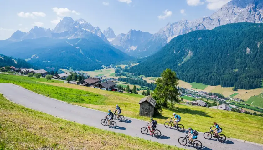 A group enjoys a cycling holiday in the Dolomites, riding down a scenic road with stunning mountain views and a charming village below.