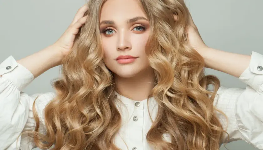 Blondynka z burzą falujących włosów, które przypominają loki na pasek od szlafroka, w białej koszuli.