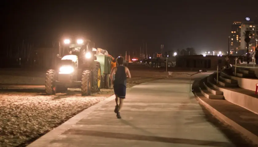Mężczyzna uprawia bieganie w nocy na promenadzie, obok pracującego traktora na plaży.