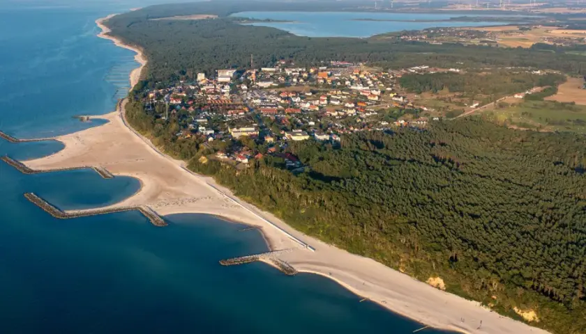 Jarosławiec atrakcje: aerial view of a coastal town with sandy beaches, breakwaters, pine forests, and a lake in the distance.