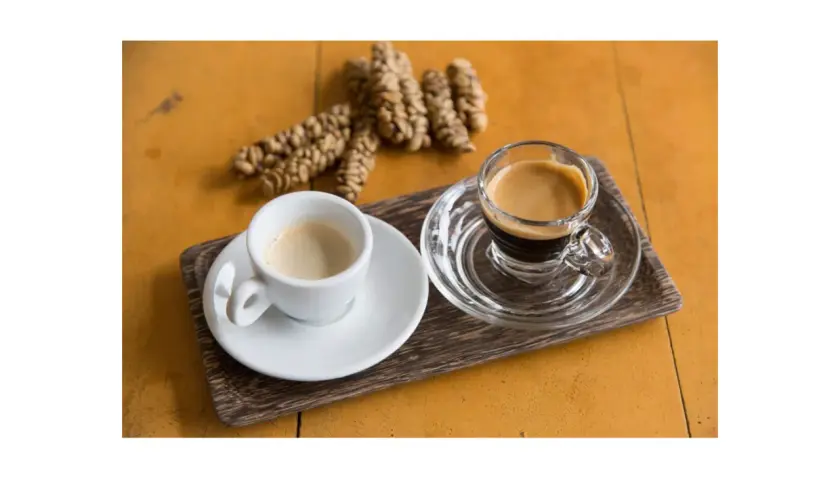Dwie filiżanki kawy, jedna biała, druga szklana, obok garść ziaren kawy. To kawa z odchodów nietoperza, znana jako Kopi Luwak.