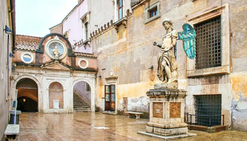 Intérieur du **château Saint-Ange** : une cour pavée et humide, une statue d'ange imposante, une arche menant à un escalier et des fenêtres grillagées.
