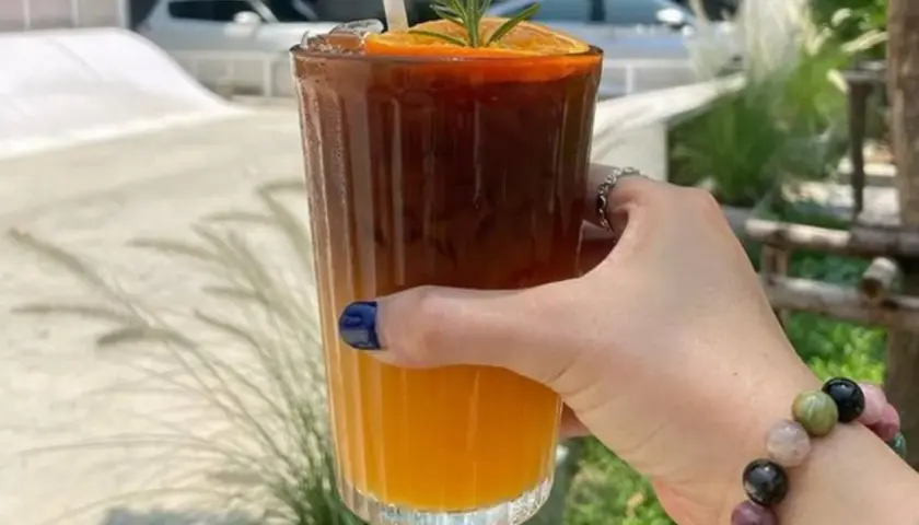 Orzeźwiające espresso z sokiem pomarańczowym, ozdobione plasterkiem pomarańczy i gałązką rozmarynu, w dłoni z kolorową bransoletką.