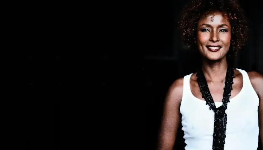 Waris Dirie w białej koszulce i czarnym naszyjniku, uśmiecha się.