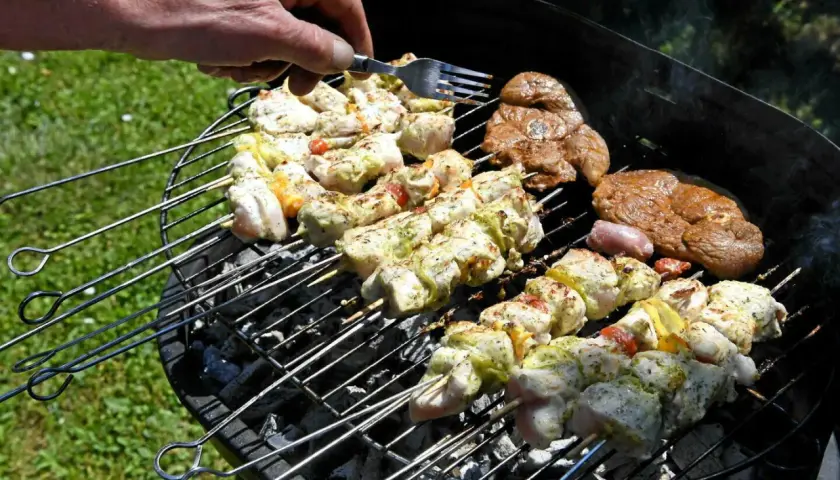 Une main vérifie des brochettes de poulet et des steaks sur un barbecue rustique.