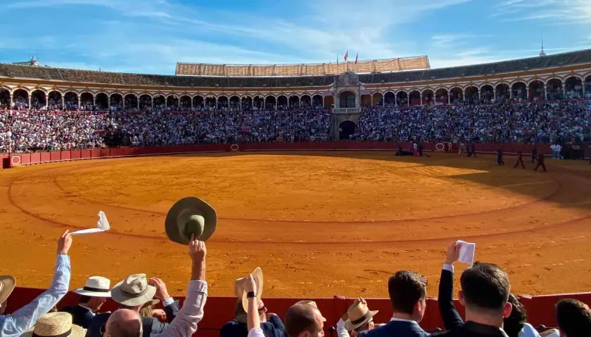 Espectadores en una **plaça de toros** levantan pañuelos y sombreros ante la arena dorada y el público en las gradas.