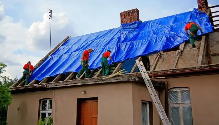 Pracownicy zabezpieczają dach niebieską plandeką. Remont dachu wymaga pozwolenia na budowę? Sprawdź przepisy.