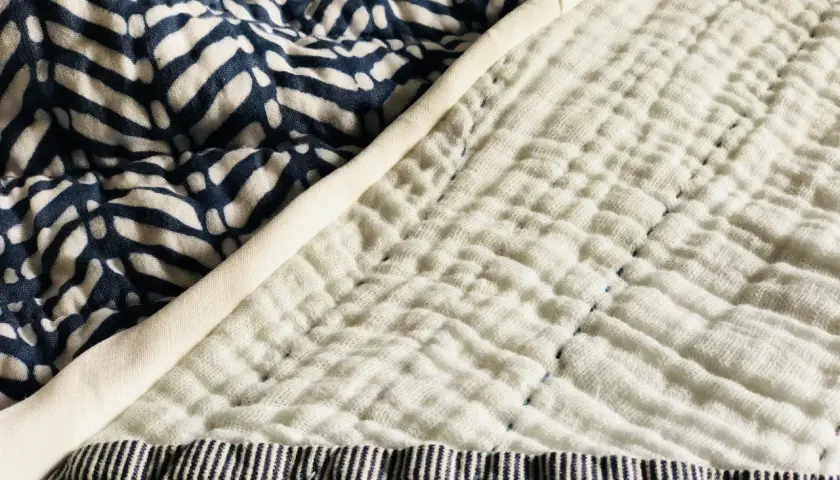 A close-up of a quilt shows a patterned blue fabric next to a textured white double gauze fabric, stitched with blue thread.