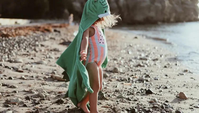 Liten badcape baby med dinosauriehuvud på stranden.