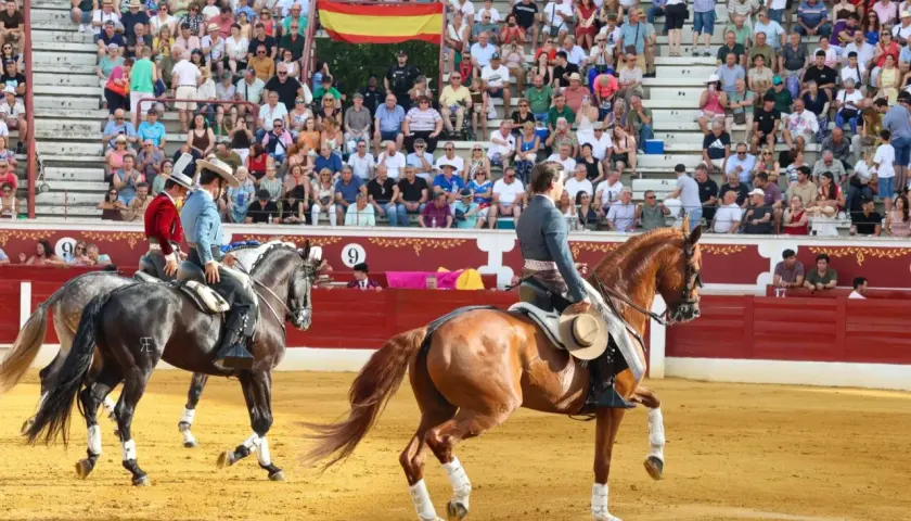 Dos jinetes, uno con chaquetilla roja y otro con azul, montan caballos en una plaza de toros. Bernardino Piriz, con sombrero, observa la escena.