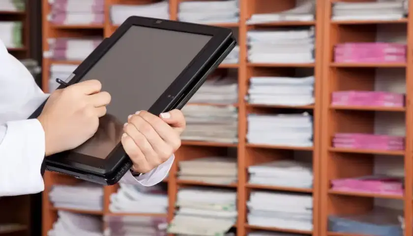 Une personne utilise une tablette pour gérer des documents, illustrant l'archivage électronique, une obligation légale. Les étagères remplies de dossiers rappellent l'importance de la numérisation.