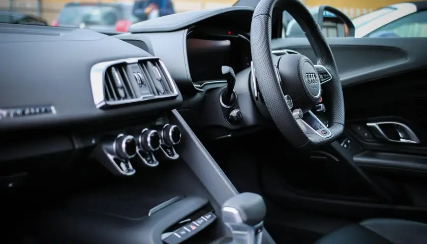 Interno di un'auto sportiva con volante a sinistra. Il cruscotto è scuro, con bocchette d'aria cromate e un display digitale. La leva del cambio è in primo piano. Perfetta per esplorare paesi con guida a sinistra.