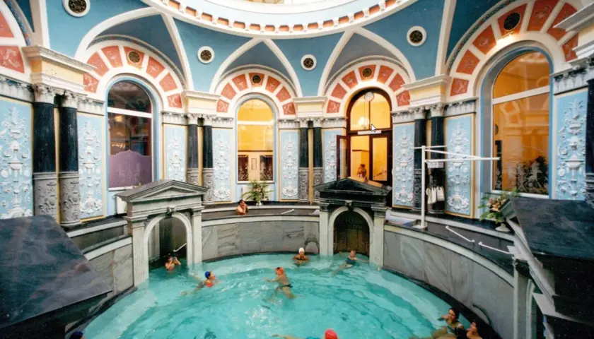 Wnętrze z basenem, gdzie ludzie relaksują się w gorącym źródle. Architektura jest bogato zdobiona, z niebieskimi i pomarańczowymi detalami.