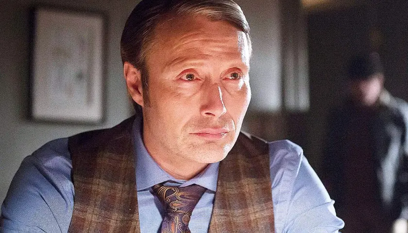 Mads Mikkelsen i en Mads Mikkelsen serie, klädd i skjorta, väst och slips. En annan person syns suddigt i bakgrunden.