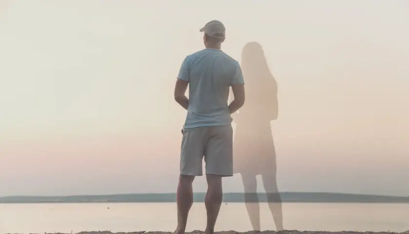 Uomo in piedi sulla spiaggia, con la figura evanescente di una donna accanto. Un'immagine che evoca il sentirsi soli in coppia.