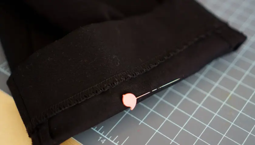 Close-up of black pants hem, pinned and ready for an invisible stitch. A pink pin secures the fabric on a cutting mat.