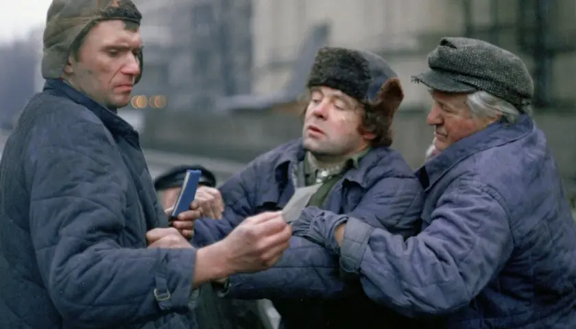 Trzech mężczyzn w zimowych kurtkach i czapkach, w tym jedna z futrzanymi uszami, dyskutuje, trzymając w rękach dokumenty. Scena z filmu "Miś" z niezapomnianą obsadą.