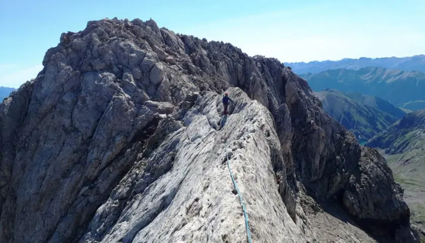 Alpinista en la cresta rocosa del Pico de Vallibierna, con un arnés y cuerda azul. Paisaje montañoso al fondo.