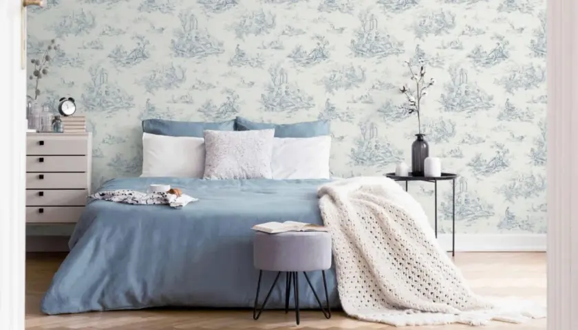 Chambre paisible avec un papier peint au motif jouy bleu et blanc, un lit bleu, des coussins blancs et une couverture en maille.