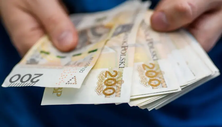 Dłonie trzymają plik banknotów 200 zł. Pytanie brzmi, po jakim czasie od zarejestrowania wypłacany jest zasiłek dla bezrobotnych.