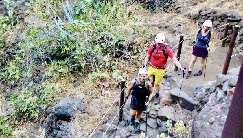 Wanderer steigen die felsige Masca Schlucht hinauf. Sie tragen Helme und Wanderstöcke.