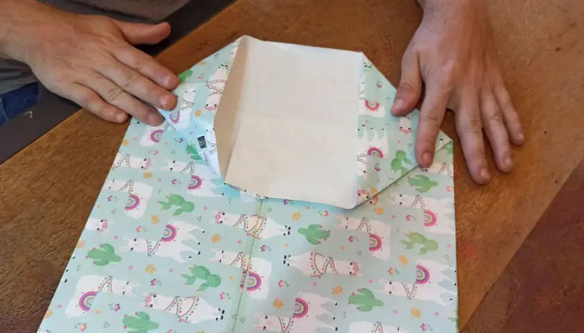 Mani che piegano carta regalo con lama e cactus per creare mini sacchetti di carta fai da te.