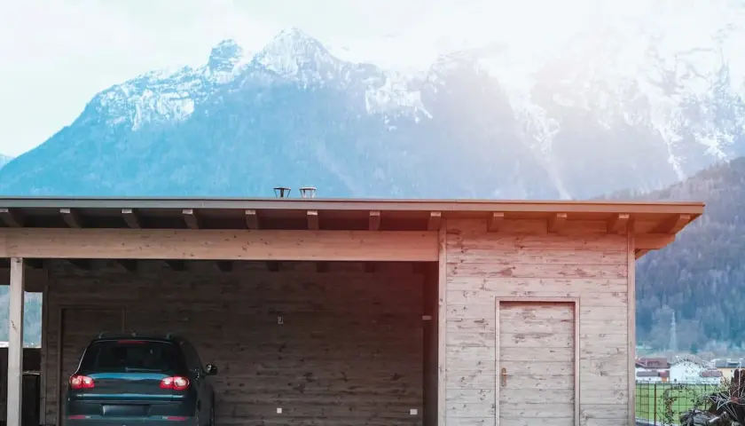 Nybyggd carport mot befintligt garage, med en bil parkerad under taket. Majestätiska berg i bakgrunden.