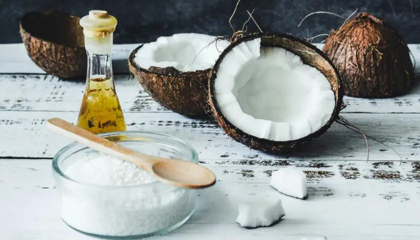 Coupé en deux, un coco frais et sa chair blanche côtoient une petite fiole d'huile dorée. Un bol de noix de coco râpée et une cuillère en bois suggèrent l'utilisation de l'huile de coco pour des recettes gourmandes ou des soins.