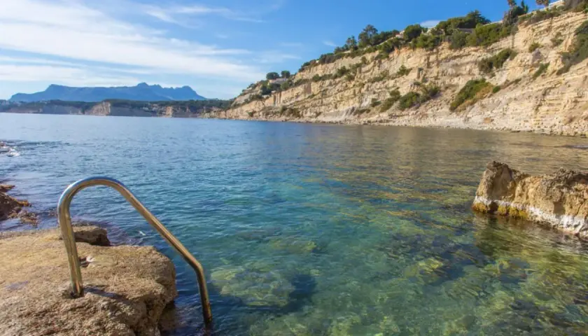 Escalera metálica para acceder a las aguas cristalinas de cala Punta Estrella, con acantilados y montañas al fondo.