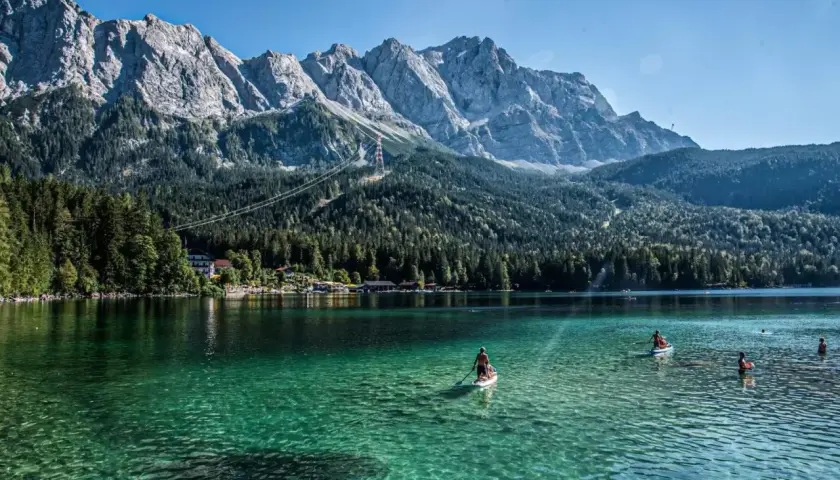 Ein Mann paddelt auf einem SUP-Board auf dem kristallklaren, blauen See in Bayern. Im Hintergrund ragen majestätische Berge auf.