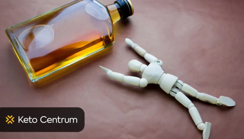 Drewniany ludzik leży obok butelki alkoholu, symbolizując negatywny wpływ alkoholu na testosteron.