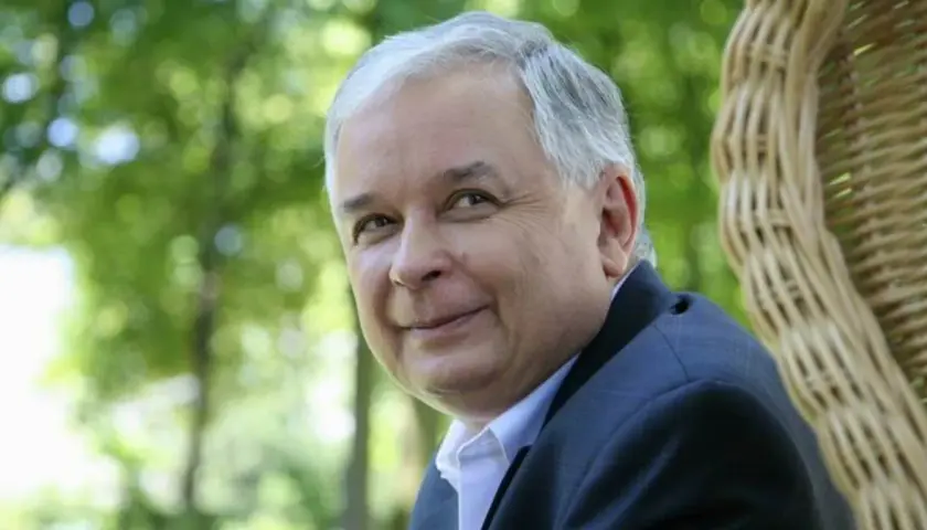 Lech Kaczyński w garniturze siedzi na tle zieleni i wiklinowego fotela.