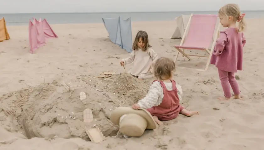 Dzieci budują zamek z piasku, ciesząc się beztroskimi zabawami na plaży. W tle widać leżaki i parawany.
