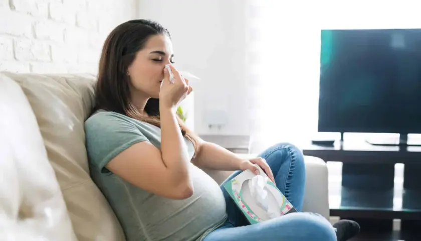 Femme enceinte s'essuyant le nez avec un mouchoir, inquiète d'un rhume enceinte danger pour bébé. Elle est assise sur un canapé, une boîte de mouchoirs à portée de main.