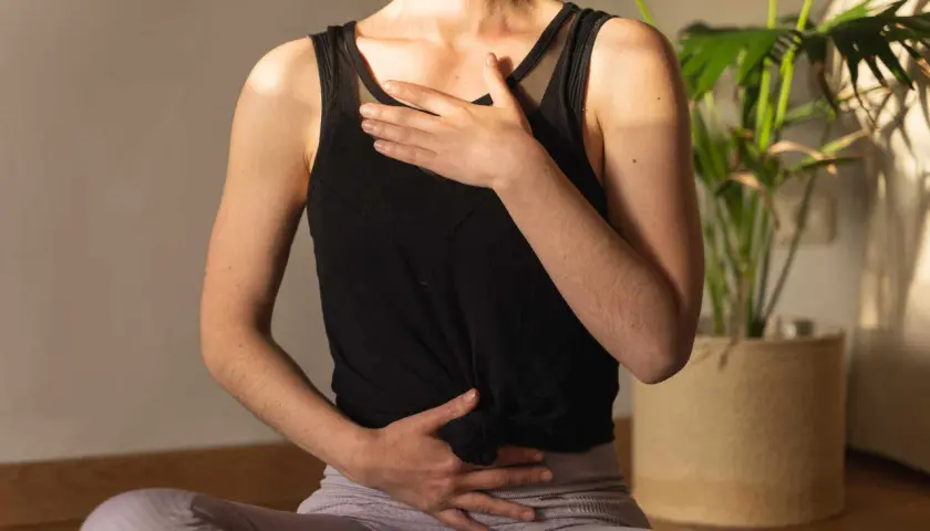 Une femme assise en tailleur, mains sur le ventre et la poitrine, pratique la respiration profonde. Une plante verte est visible en arrière-plan.