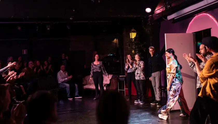 Publik applåderar en föreställning av improvisationsteater i Stockholm. Skådespelare på scenen tar emot hyllningar.