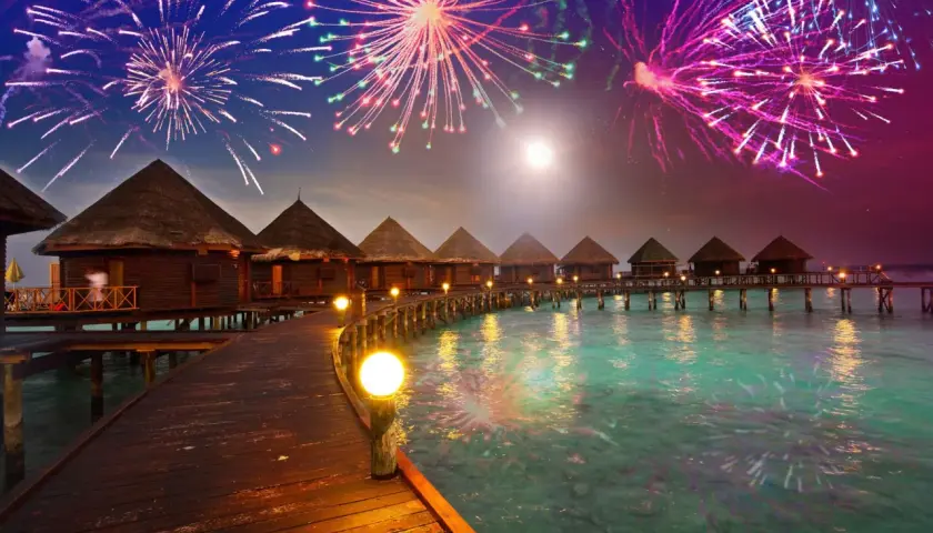 Bungalows auf Stelzen über türkisfarbenem Wasser, beleuchteter Steg, Feuerwerk am Nachthimmel. Hier ist es an Silvester warm und feierlich.