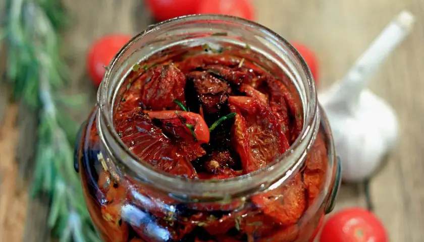 Pomodori secchi sott'olio, la ricetta della nonna. Un barattolo pieno di sapore, con rosmarino e aglio sullo sfondo.