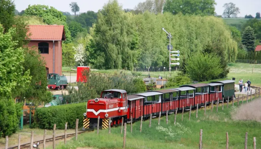 Czerwony pociąg z wagonami turystycznymi jedzie przez zielone tereny. W tle budynek i drzewa. To świetne pałuki atrakcje dla całej rodziny.