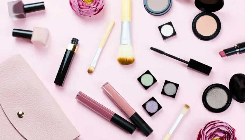 En rosa bakgrund med olika sminkprodukter som läppglans, mascara, puder och borstar. Perfekt smink i handbagage för en snabb touch-up.