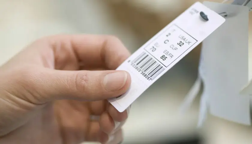 Main d'une personne tenant une étiquette de vêtement avec un code-barres et des tailles.