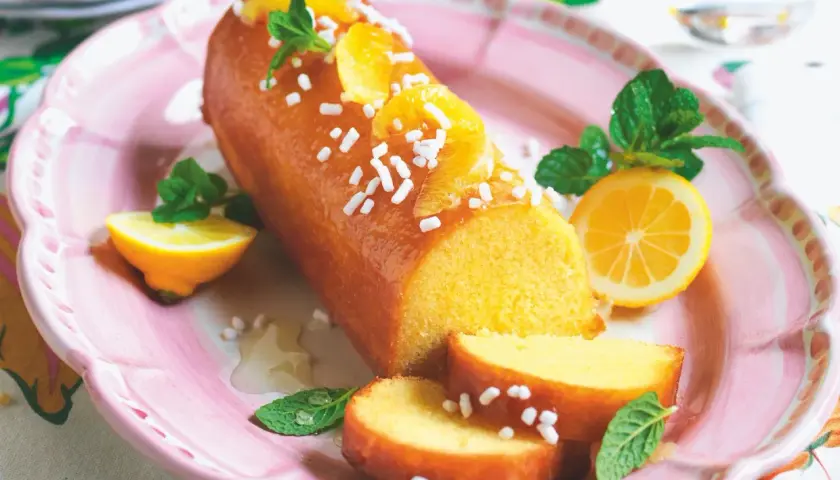 Una deliziosa torta al limone glassata, decorata con fette d'arancia, zuccherini e menta, servita su un piatto rosa con spicchi di limone.