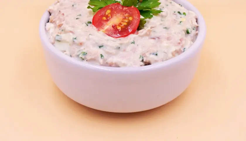 Pyszna pasta z tuńczyka i jajka, udekorowana pomidorkiem i natką pietruszki, w białej miseczce.