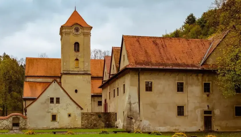 Czerwony klasztor zaprasza na zwiedzanie. Zabytkowa wieża i budynki z pomarańczowymi dachami otoczone zielenią.