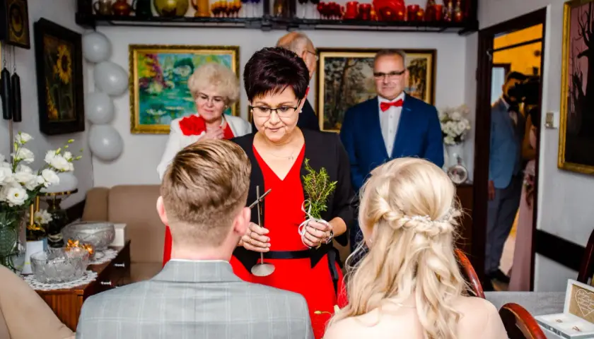 Kobieta w czerwonej sukience udziela nowoczesne błogosławieństwo parze młodej, trzymając gałązkę.