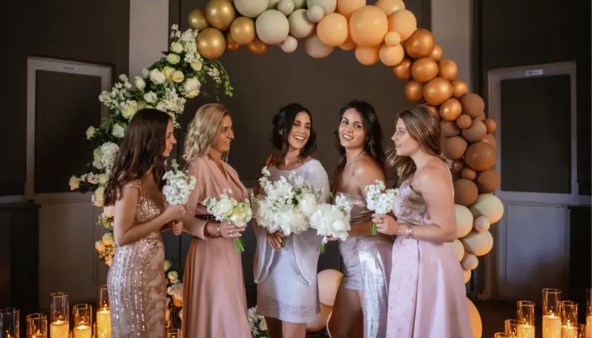 Cinq femmes souriantes, entourées de fleurs et de ballons, se préparent pour un mariage. Elles tiennent des bouquets, prêtes pour la cérémonie.