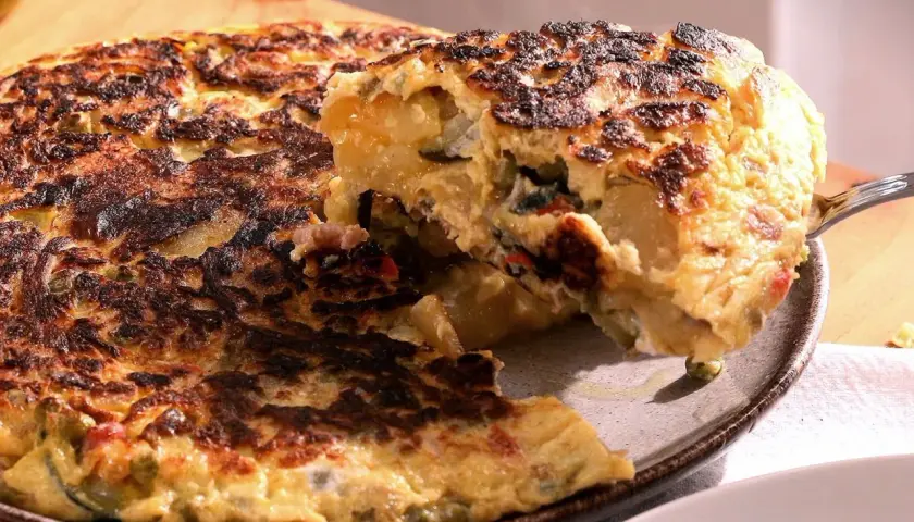 Una porción de tortilla paisana, dorada y jugosa, se levanta con un tenedor, revelando patatas, pimientos y huevo.
