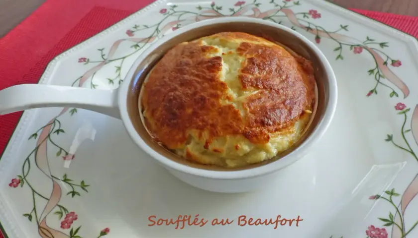 Un délicieux soufflé au beaufort, doré et gonflé, servi dans un ramequin sur une assiette décorée de rubans et de fleurs.